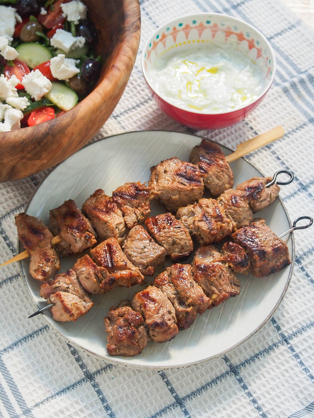 This flavorful Greek lamb marinade is easy to prepare and perfect to make lamb souvlaki skewers, or for larger chops or steaks. Delicious with pita, salad, tzatziki and more. three Greek lamb souvlaki skewers with salad and bowl of twatziki sauce behind.