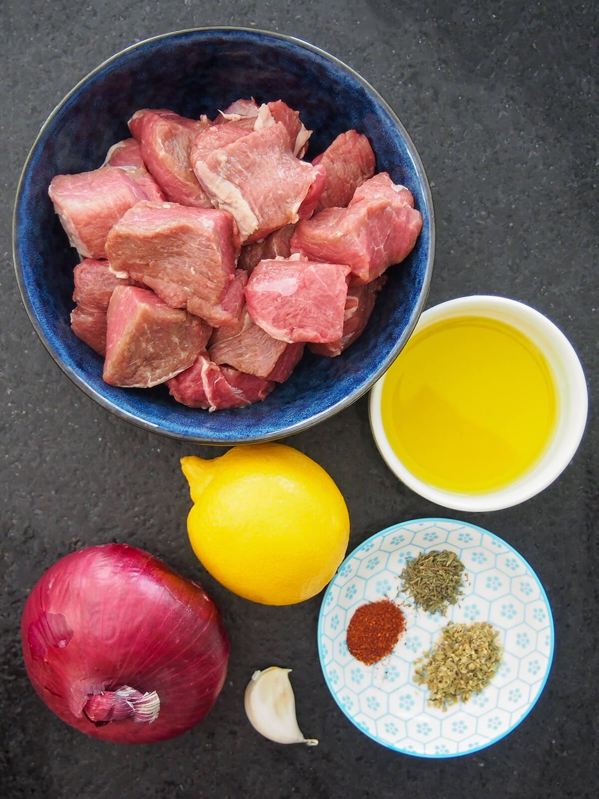 ingredients for Greek marinated lamb souvlaki bowl of diced lamb, bowl of olive oil, lemon, red onion, and plate with herbs and spices.
