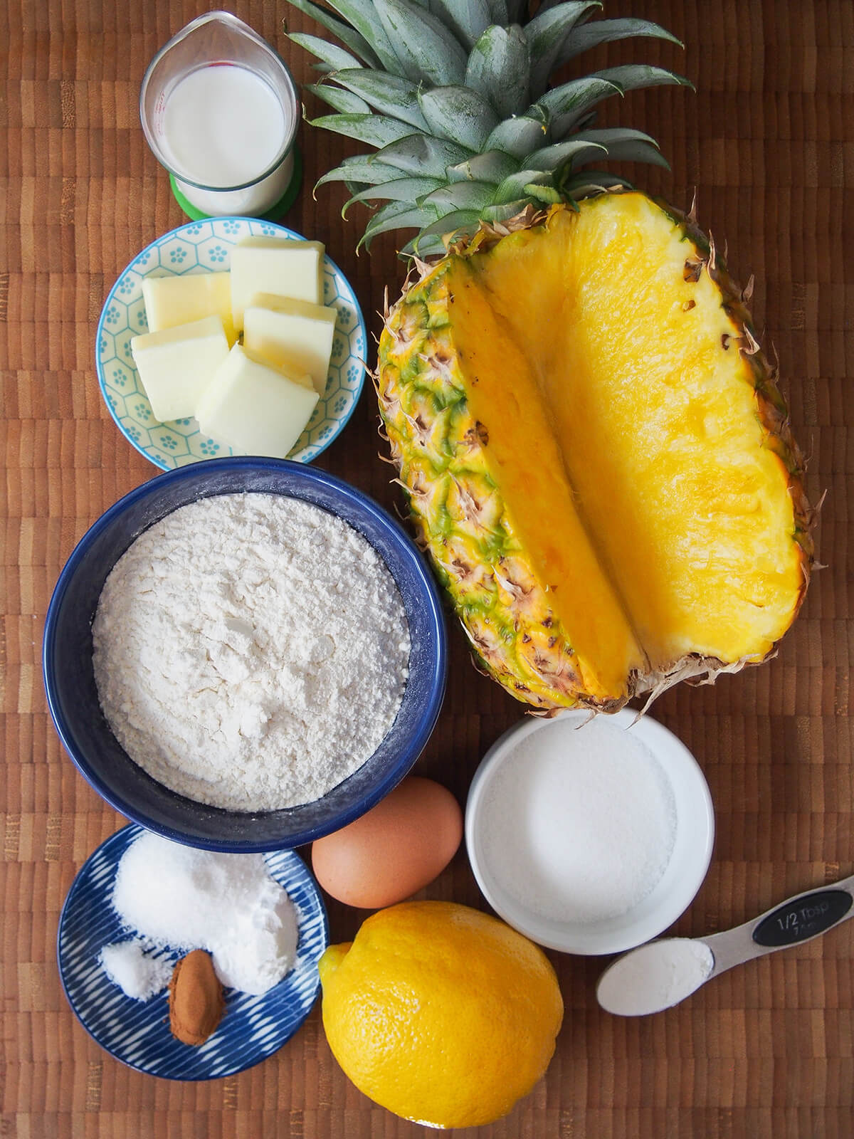 pineapple with a chunk cut out of it, bowls with flour, butter, sugar, measure of milk egg, lemon and dish with spices.