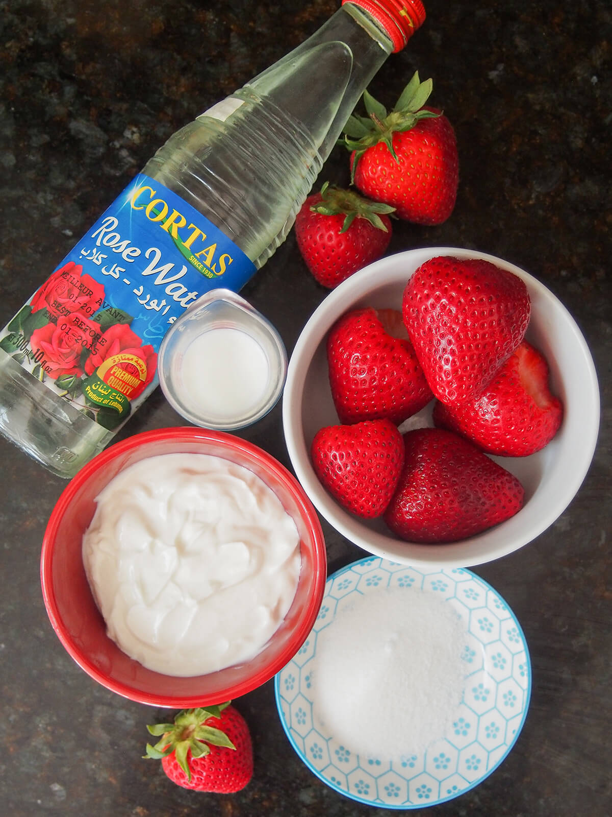ingredients for strawberry lassi bottle of rosewater, bowls of strawberries and yogurt with some milk and sugar to side.