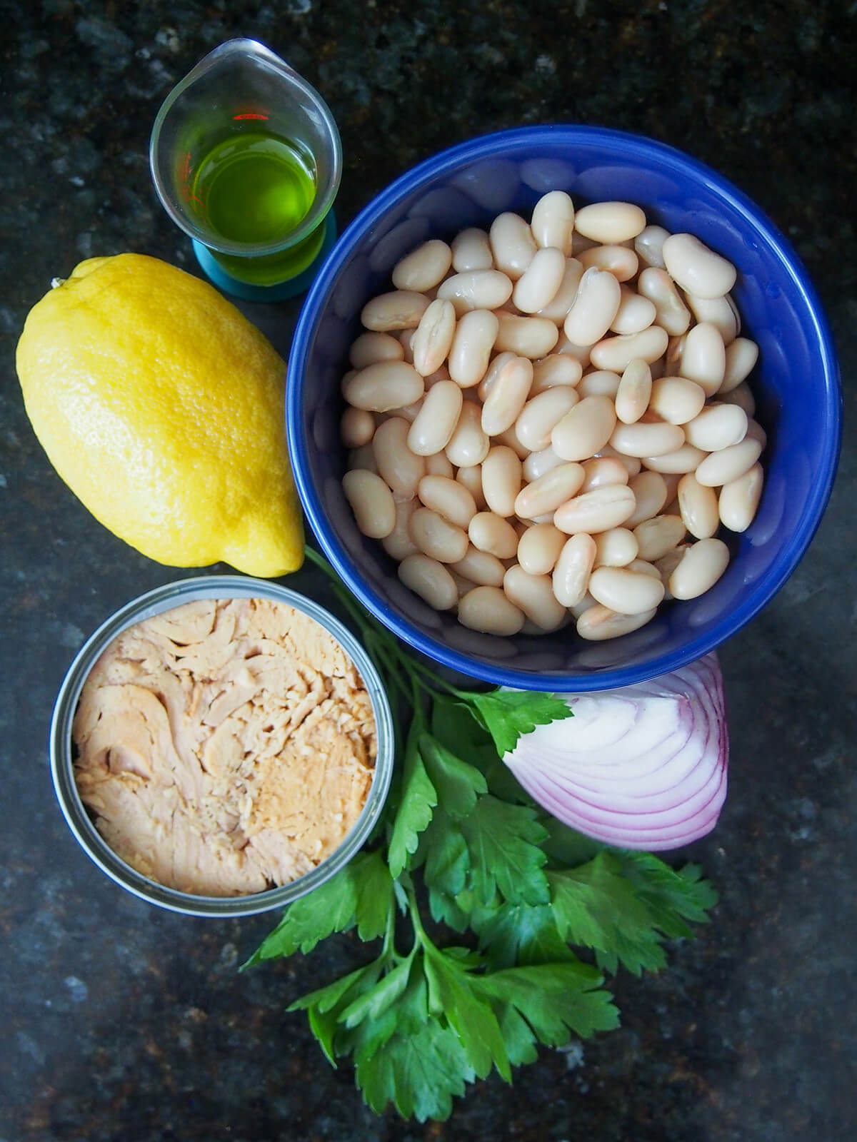 ingredients for white bean and tuna salad blue bowl filled with beans open can of tuna, parsley, lemon and part red onion.