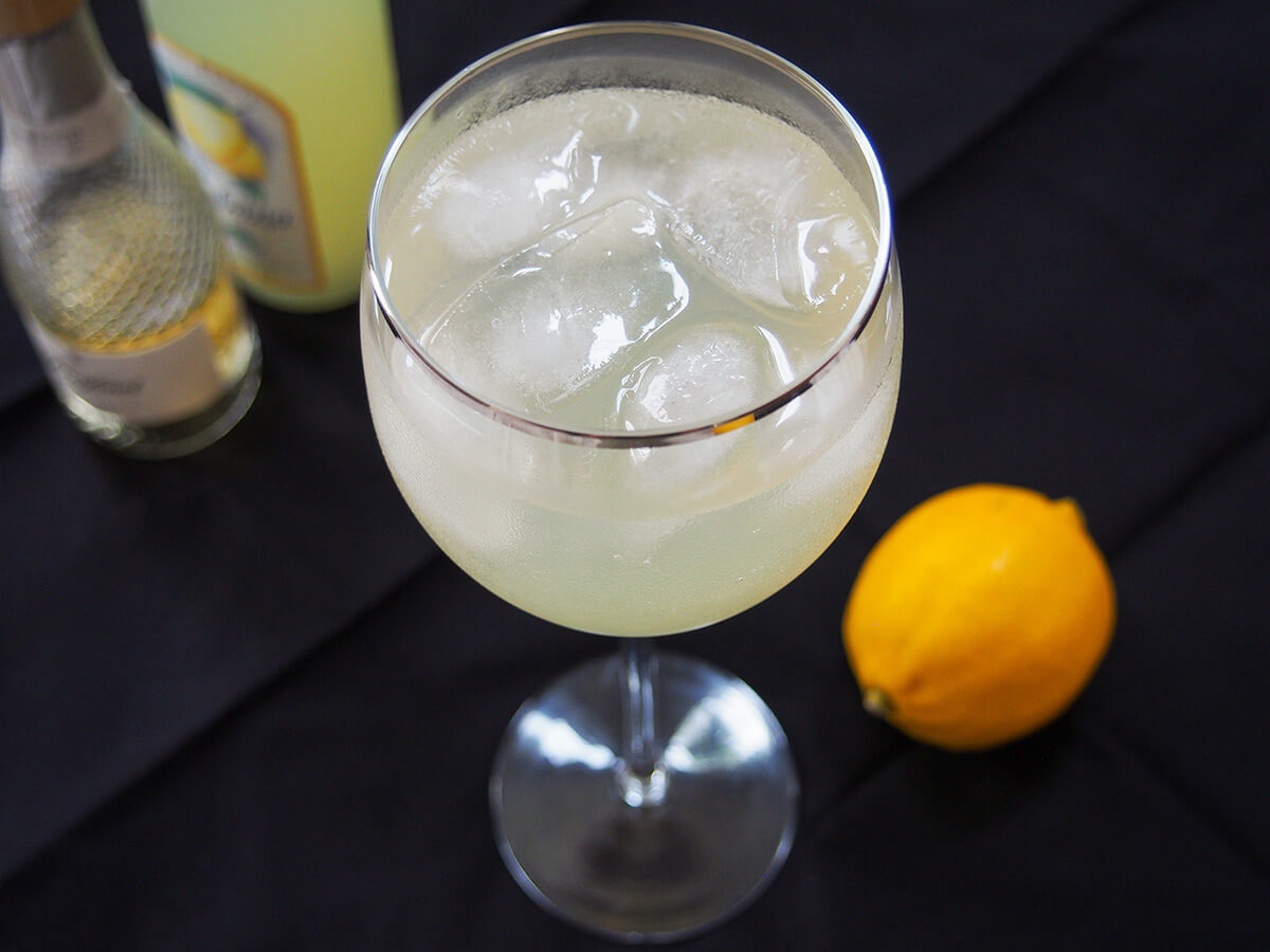 glass of limoncello spritz with lemon to right and bottles in background behind.