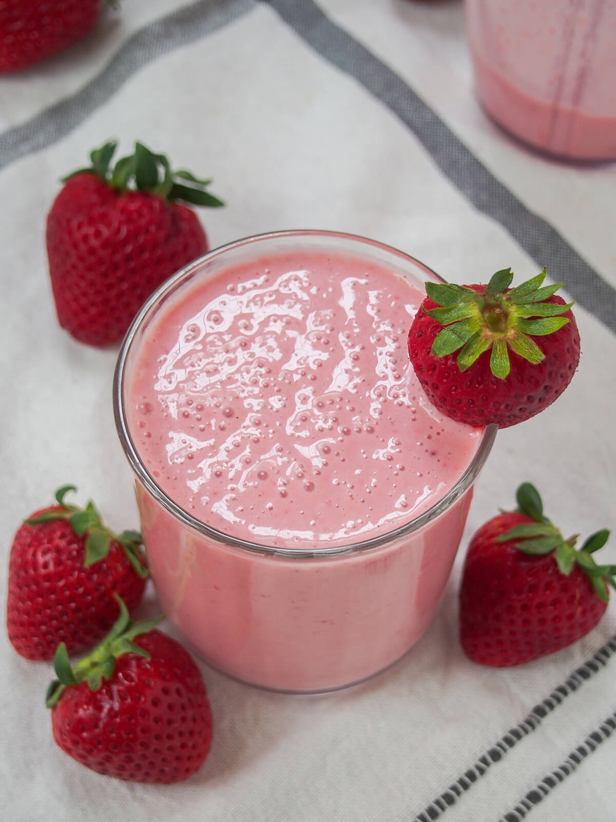 Strawberry lassi is a gently fruity and colorful twist on the classic Indian yogurt-based drink. It's kind of a light smoothie, made with just a handful of ingredients, that's smooth, creamy and refreshing. glass of strawberry lassi with strawberries around glass and bubbles on top of glass.