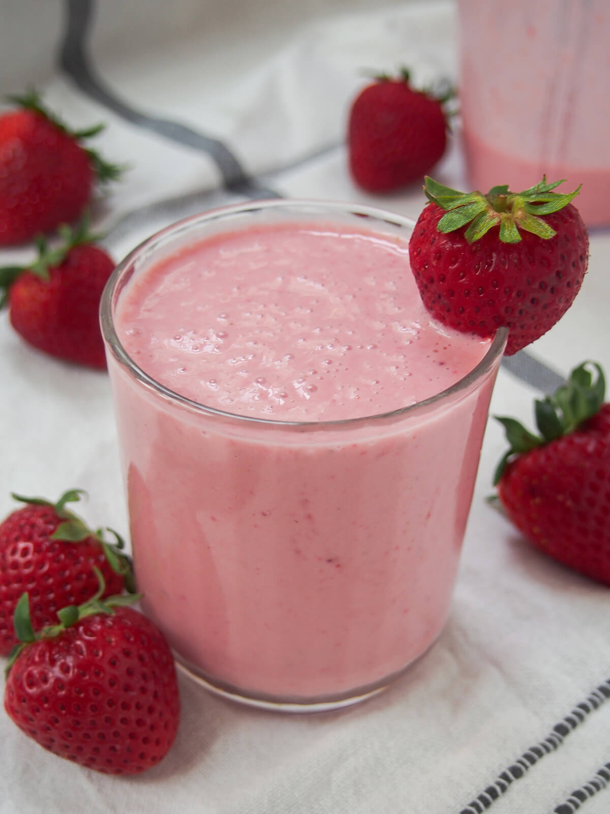 Strawberry lassi is a gently fruity and colorful twist on the classic Indian yogurt-based drink. It's kind of a light smoothie, made with just a handful of ingredients, that's smooth, creamy and refreshing. side view of glass of strawberry lassi with strawberries and blender jug with small amount left behind.