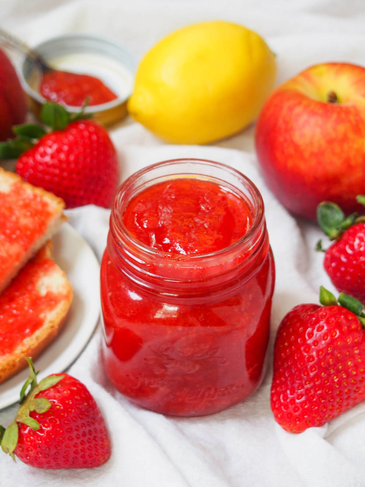 jar of strawberry peach jam with part view of peach and lemon behind, strawberries and toast to sides.