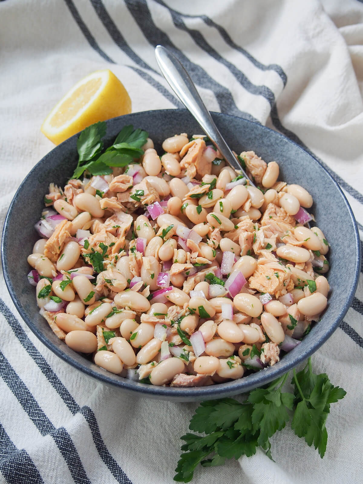 This Tuscan white bean and tuna salad (fagioli e tonno) is really easy to make with just a handful of ingredients. The result is a a tasty mixture of flavors that works well as an antipasti, potluck side or simply a quick lunch. bowl of Tuscan white bean and tuna salad with half a lemon behind bowl and parsley in front.