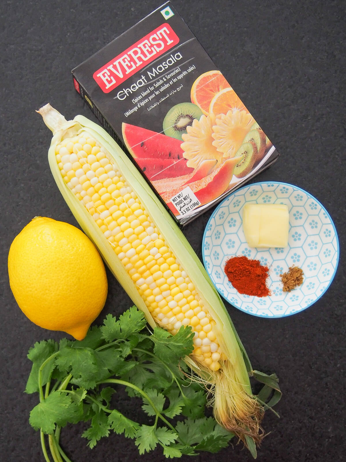 corn cob, lemon, bowl with spices and butter, bunch of cilantro and packet of chaat masala from overhead.