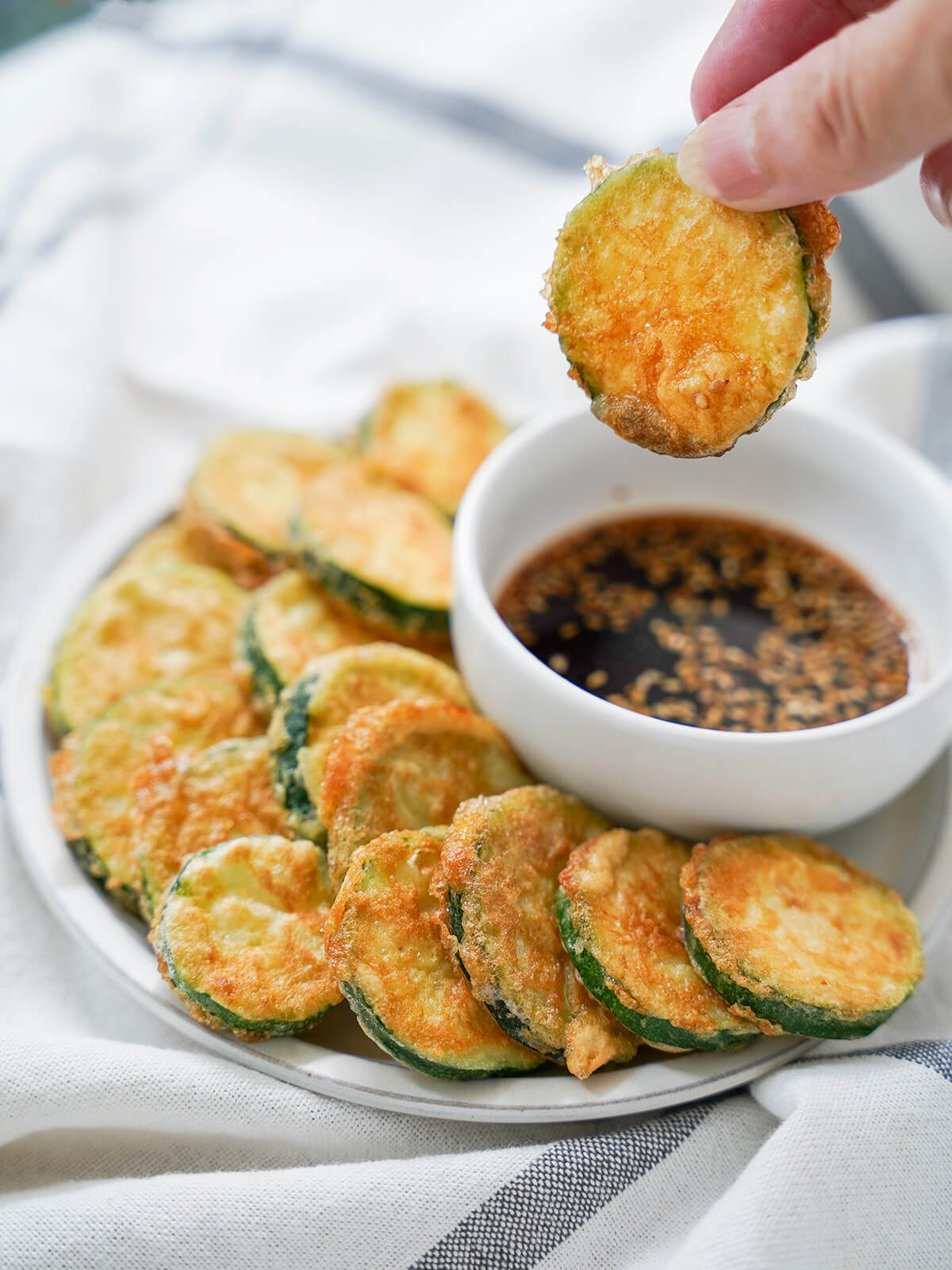holding a zucchini fritter over dipping sauce with plate of fritters below.