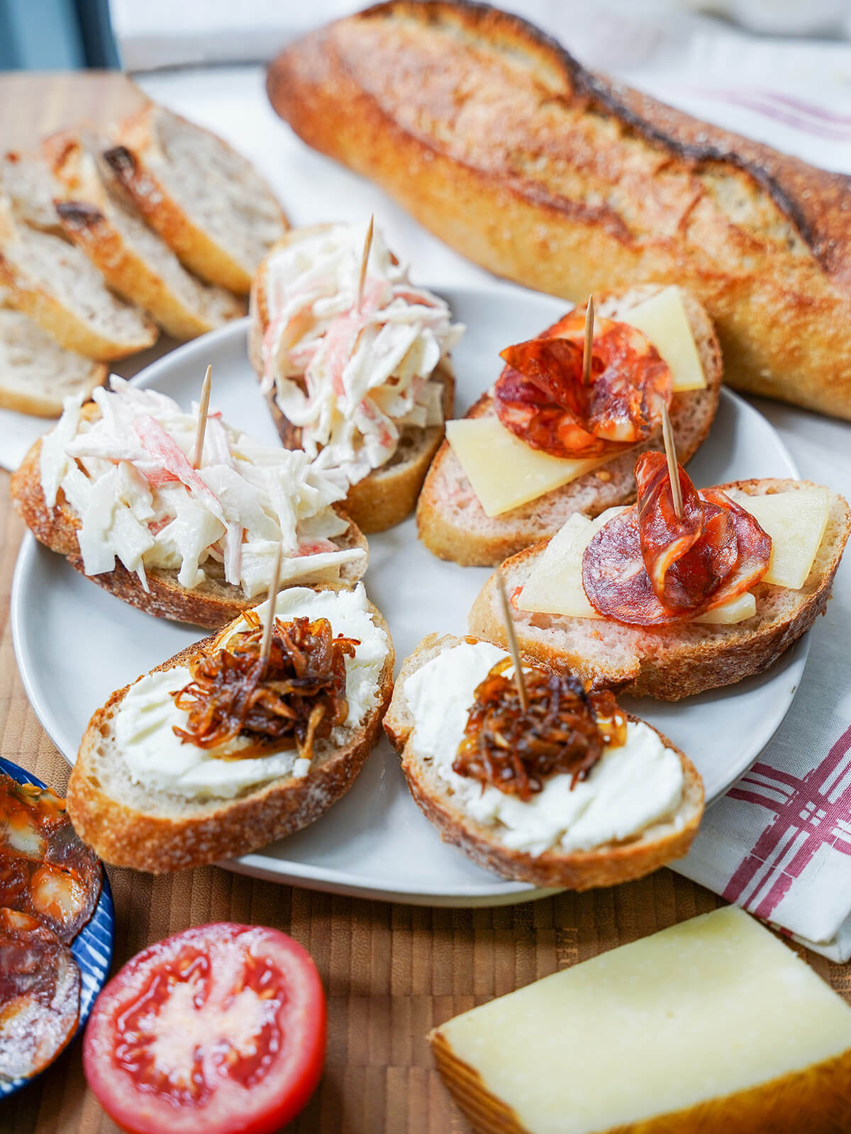 Spanish pintxos are simple finger food tapas that have lots of scope for variety. Get some inspiration, try some easy pintxos recipes and enjoy these tasty bites at home. side view of plate of Spanish pintxos with part baguette and slices behind, block of cheese in front.