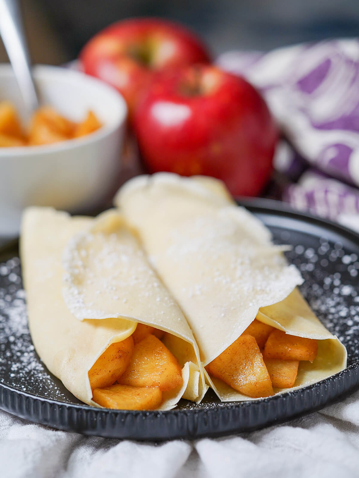side view of two apples crepes with apples and dish with caramelized apples blurred in background.