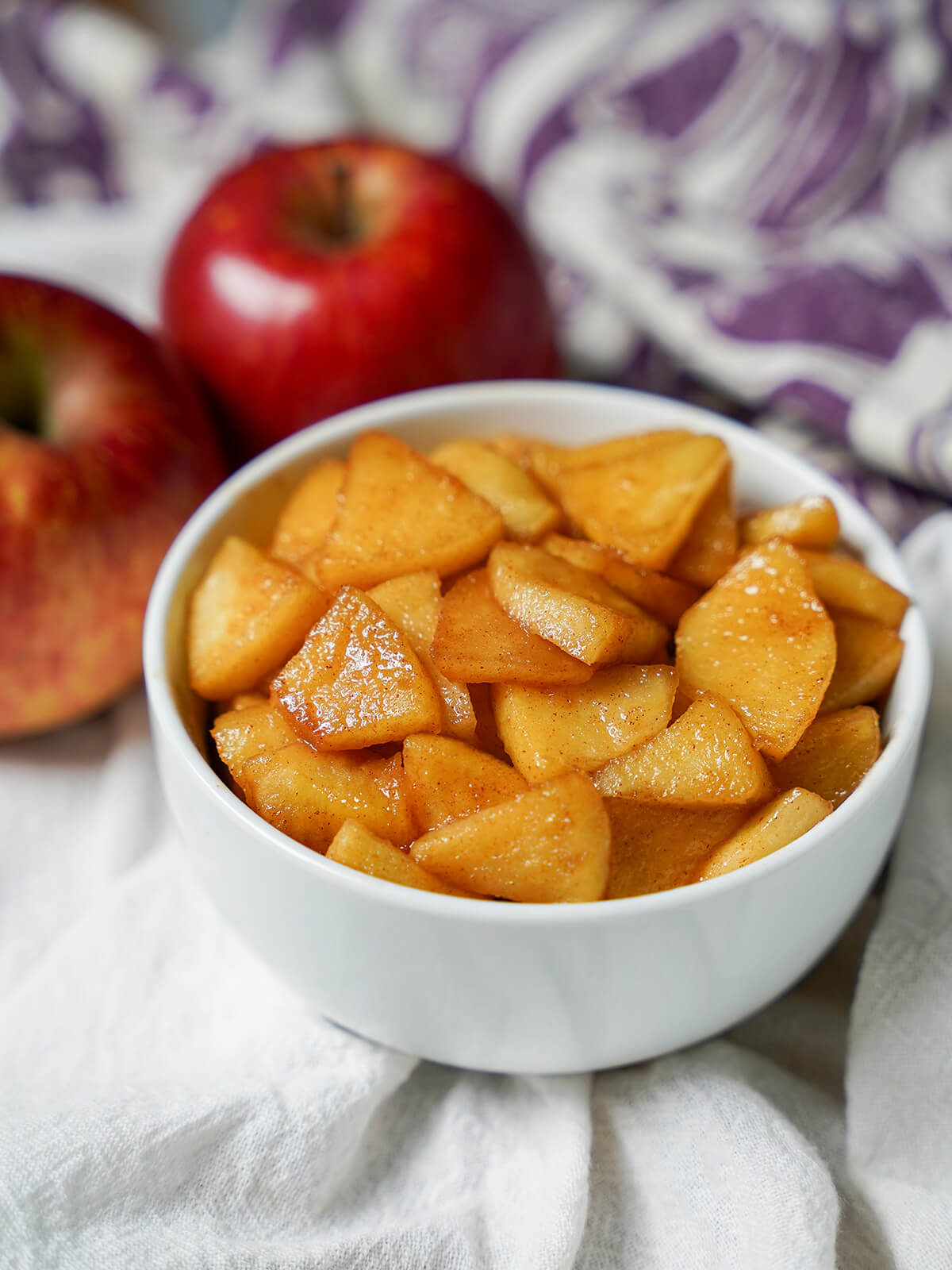 bowl of caramelized apples with apples and purple cloth in background behind.