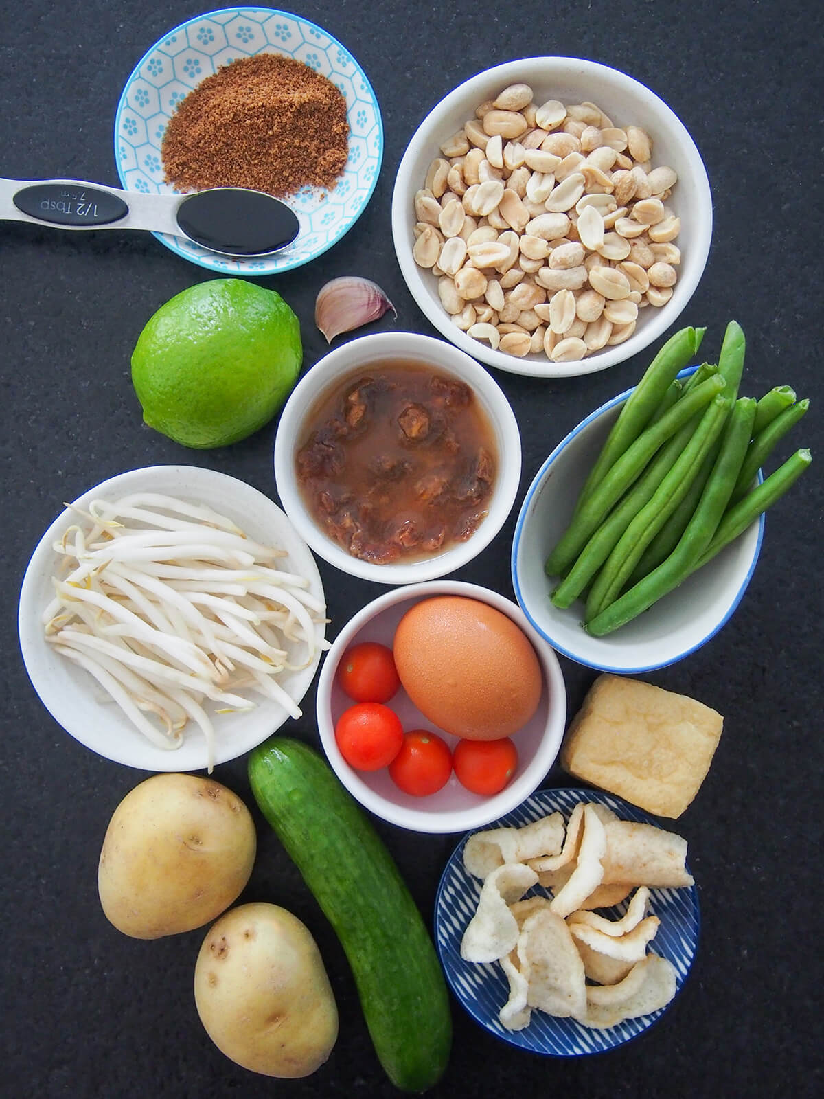 bowls of peanuts, green beans, tamarind, beansprouts, egg, crackers with cucumber, potato, tofu to side from overhead.