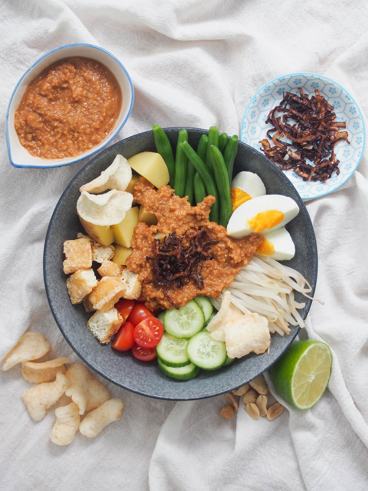 overhead view of bowl of Gado gado (Indonesian salad with peanut sauce) with sauce, shallots and crackers around bowl.