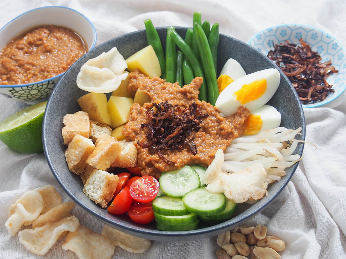 side view of bowl of Gado gado (Indonesian salad with peanut sauce) with sauce and fried shallots in dishes behind.