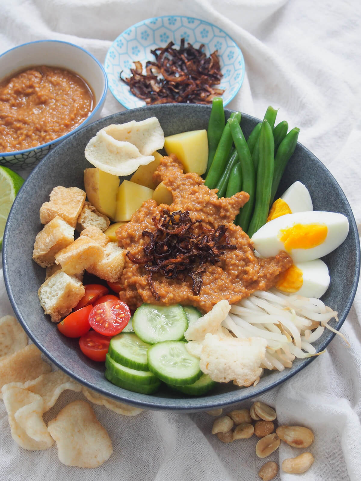 bowl of Gado gado (Indonesian salad with peanut sauce) with sauce and shallots behind.