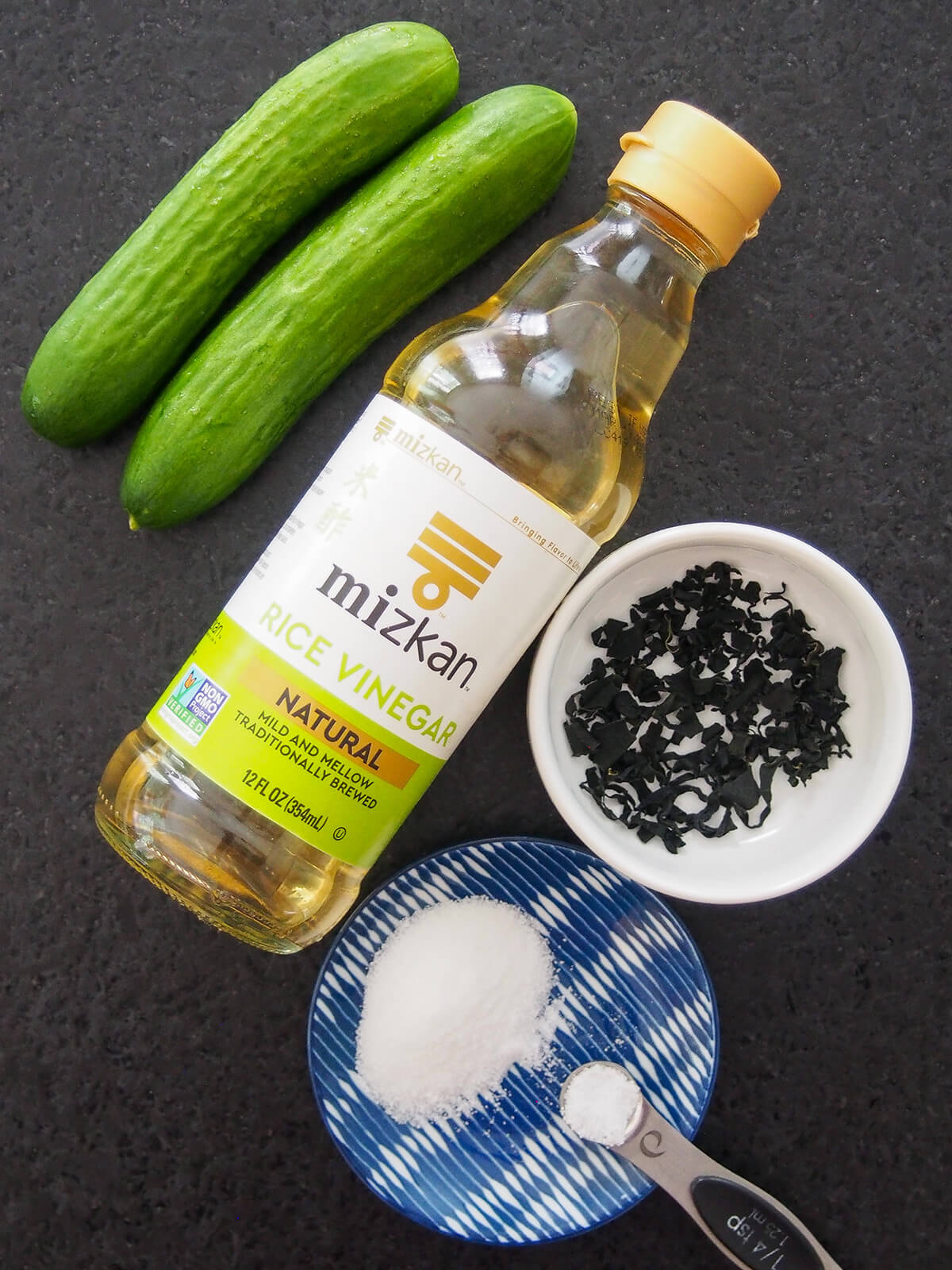 small cucumbers, bottle of rice vinegar, dishes with dried wakame and sugar and salt.