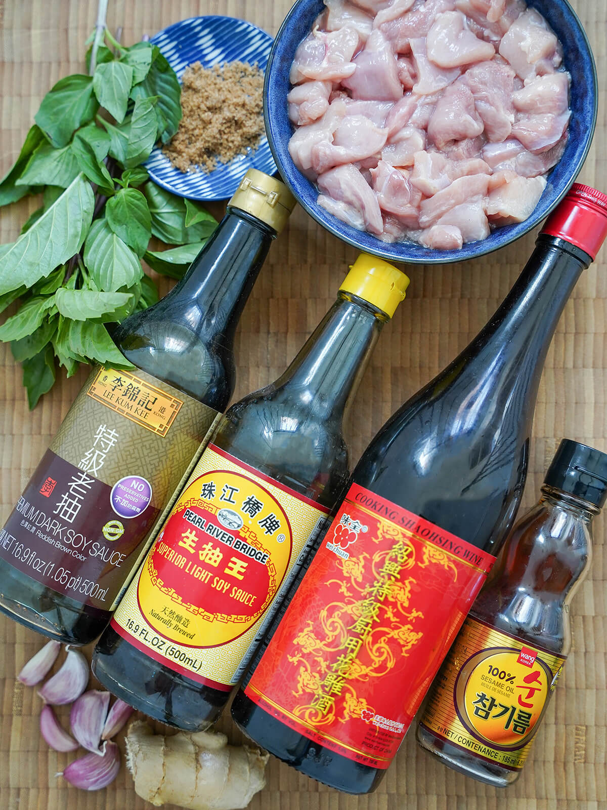 ingredients for Taiwanese three cup chicken bottles of light and dark soy sauce, shaoxing wine, sesame oil, bowl of chopped chicken, garlic cloves, ginger and Thai basil bunch.