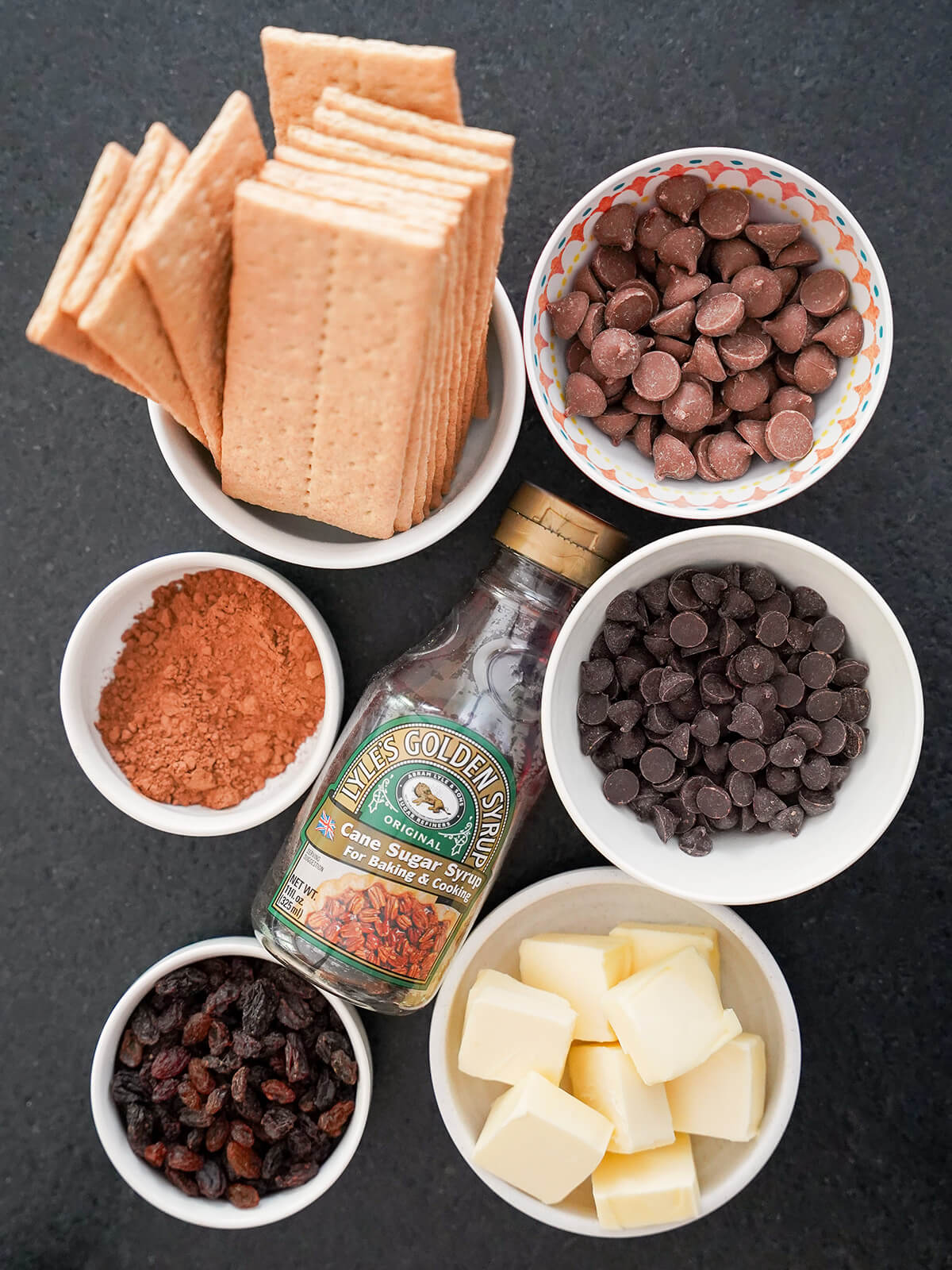 bowls of chocolate chips, butter, graham crackers, raisins and cocoa powder with bottle of golden syrup in middle.