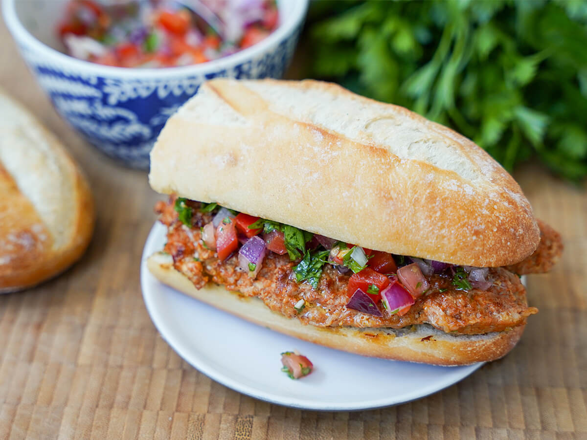 choripan, Argentine chorizo sandwich, on plate with bowl of salsa and parsley blurred in background behind.