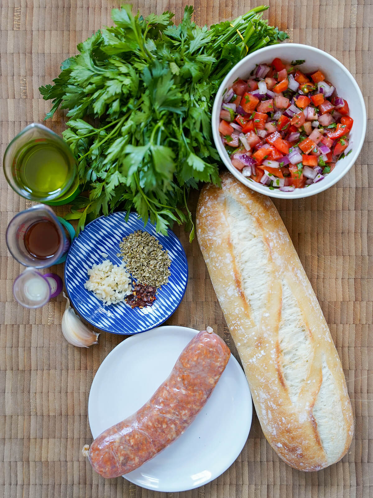 chorizo sausage, demi baguette, bowl of salsa, bunch of parsley, oil, vinegar and small plate with oregano, garlic and chili.