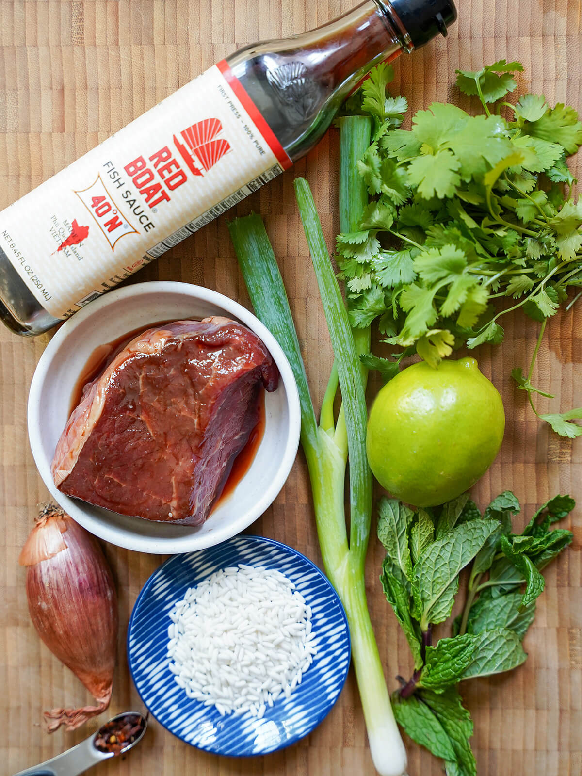 bottle of fish sauce, lime, herbs, green onion, shallot and plates with marinated beef and rice.