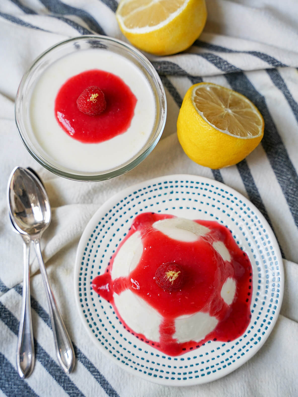 overhead view of lemon panna cotta on a plate with raspberry coulis drizzled over and another panna cotta in dish with spoons and lemon to side.
