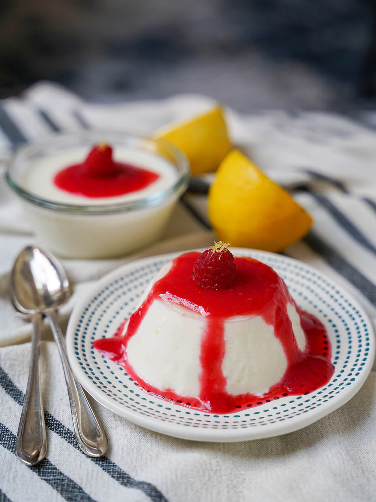 plate of lemon panna cotta with spoons to side, another panna cotta in dish behind and lemons.
