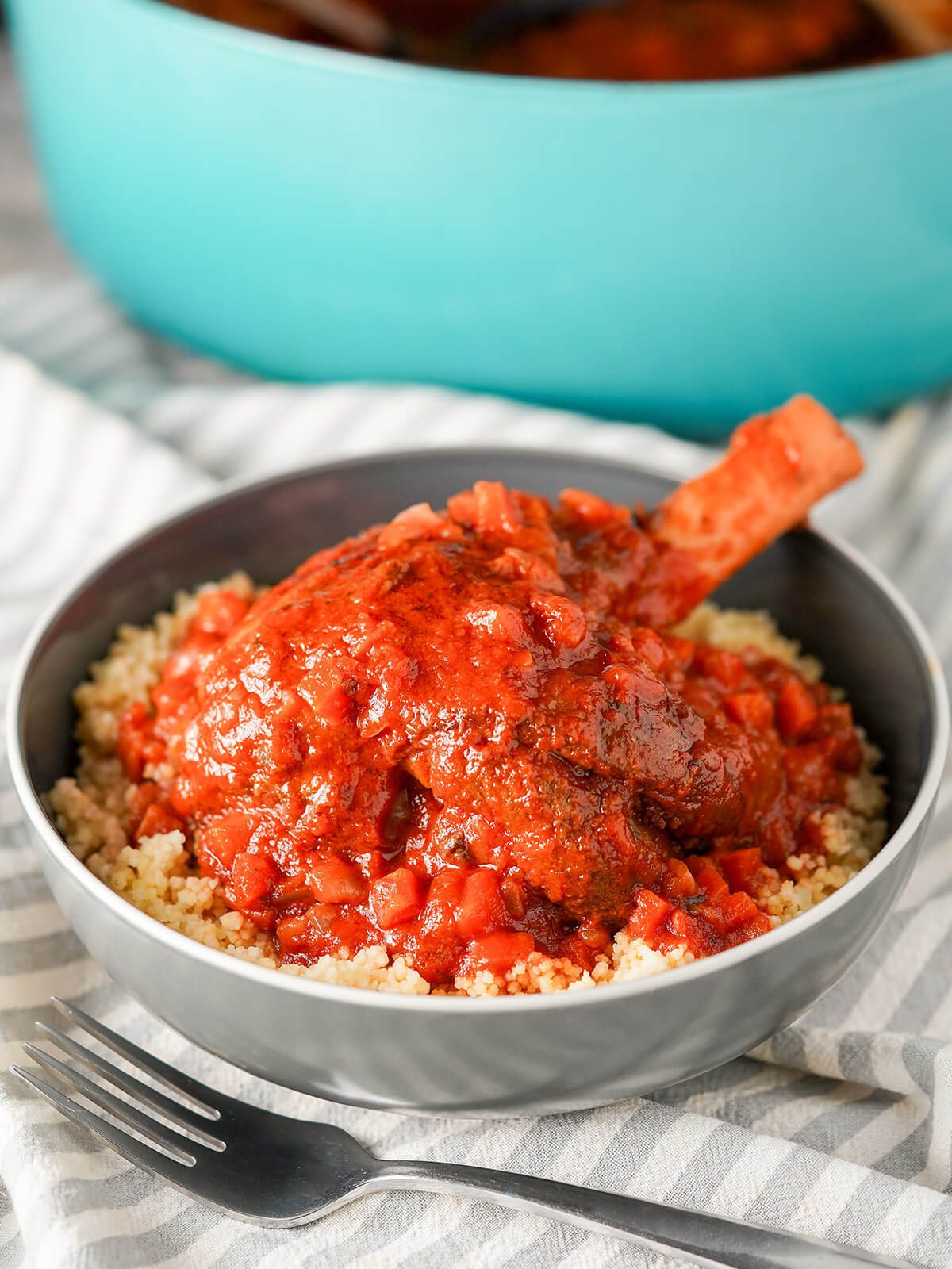 bowl of braised lamb shanks over couscous with pot in background behind.