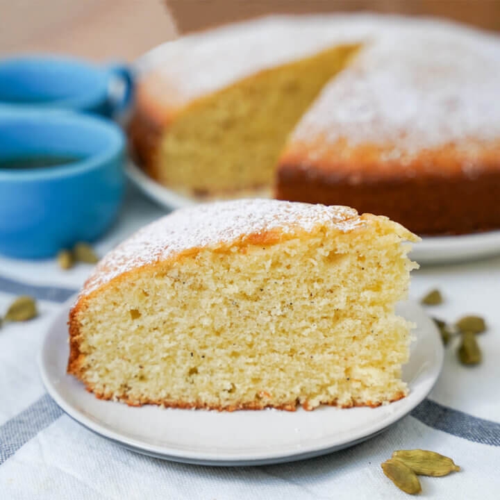 slice of cardamom cake with rest of cake and cups fuzzy in background.