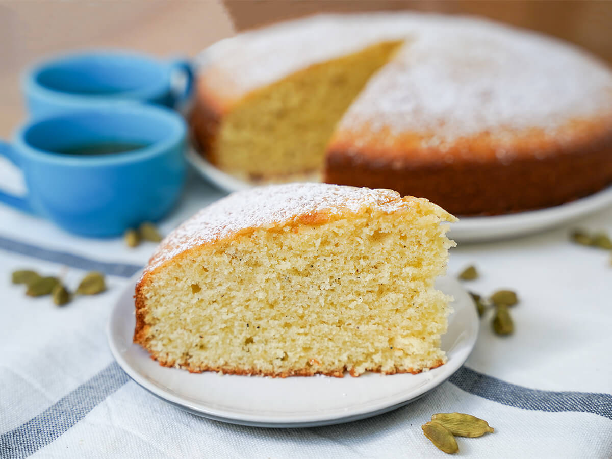 slice of cardamom cake on plate with cardamom pods either side, rest of cake and cups behind.