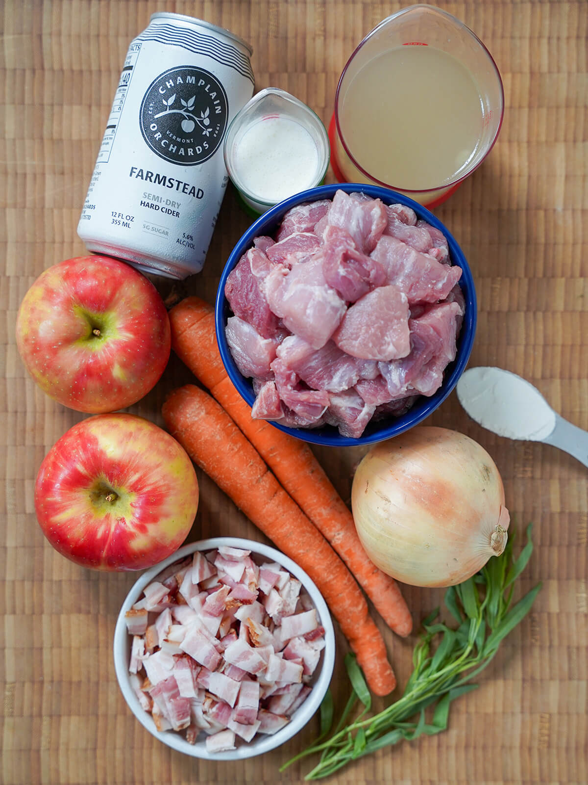 dishes of diced pork, bacon with apples, carrots, onion and can of cider around.