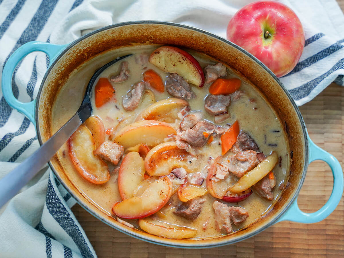 pot of pork and cider casserole with spoon to one side and apple above dish.