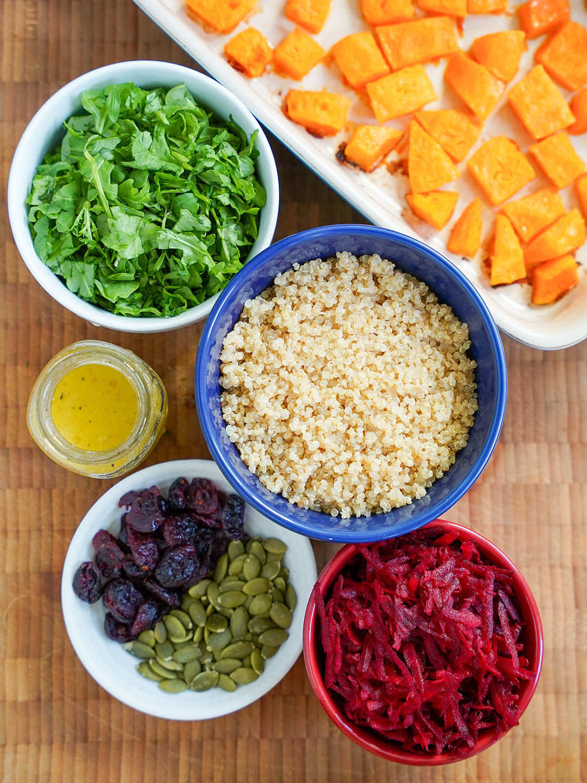 bowls of chopped arugula, grated beet, cooked quinoa, cranberries and pepitas with dish of squash to side.