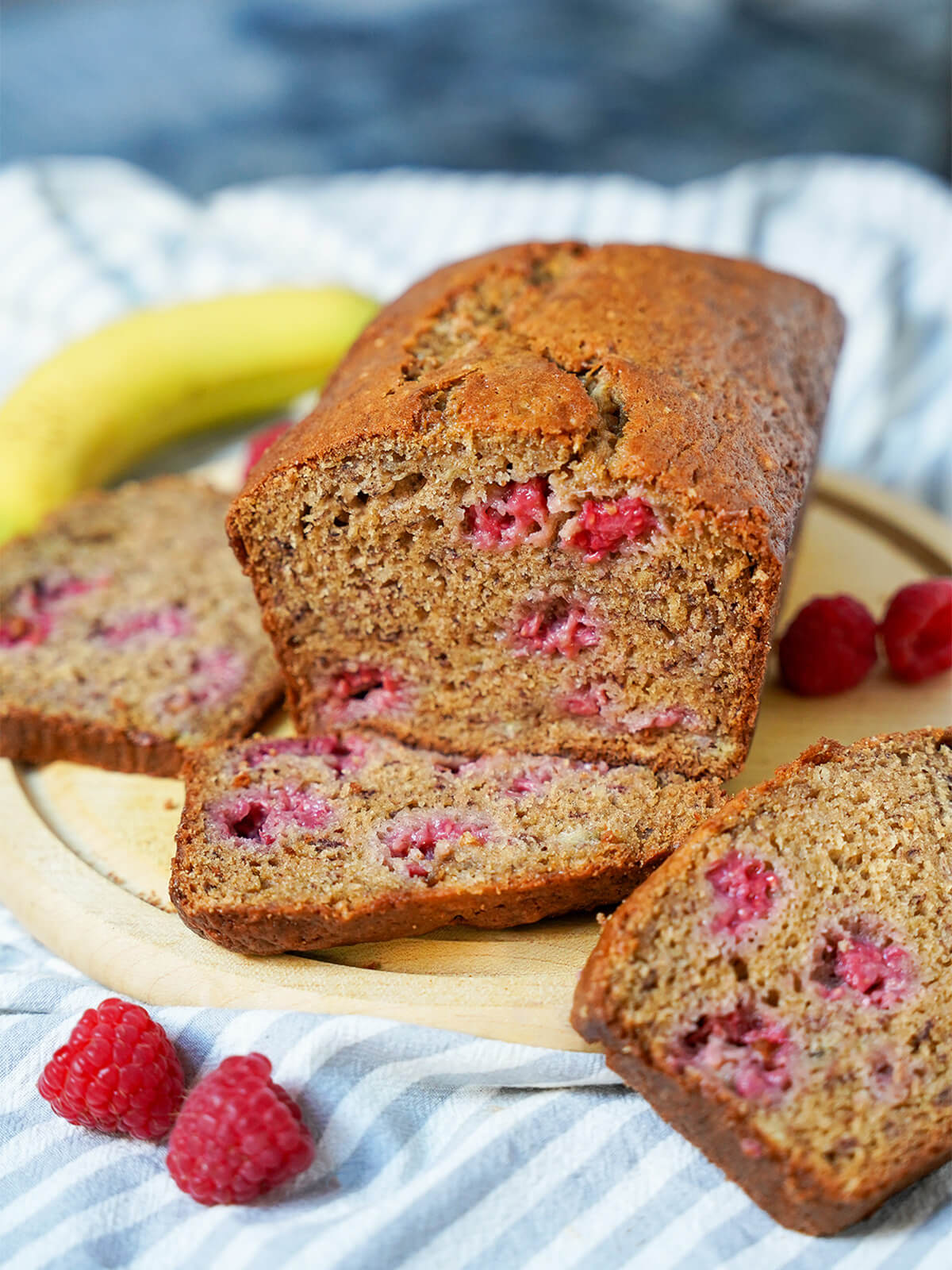 loaf of raspberry banana bread on board with slices, raspberries and banana around it.