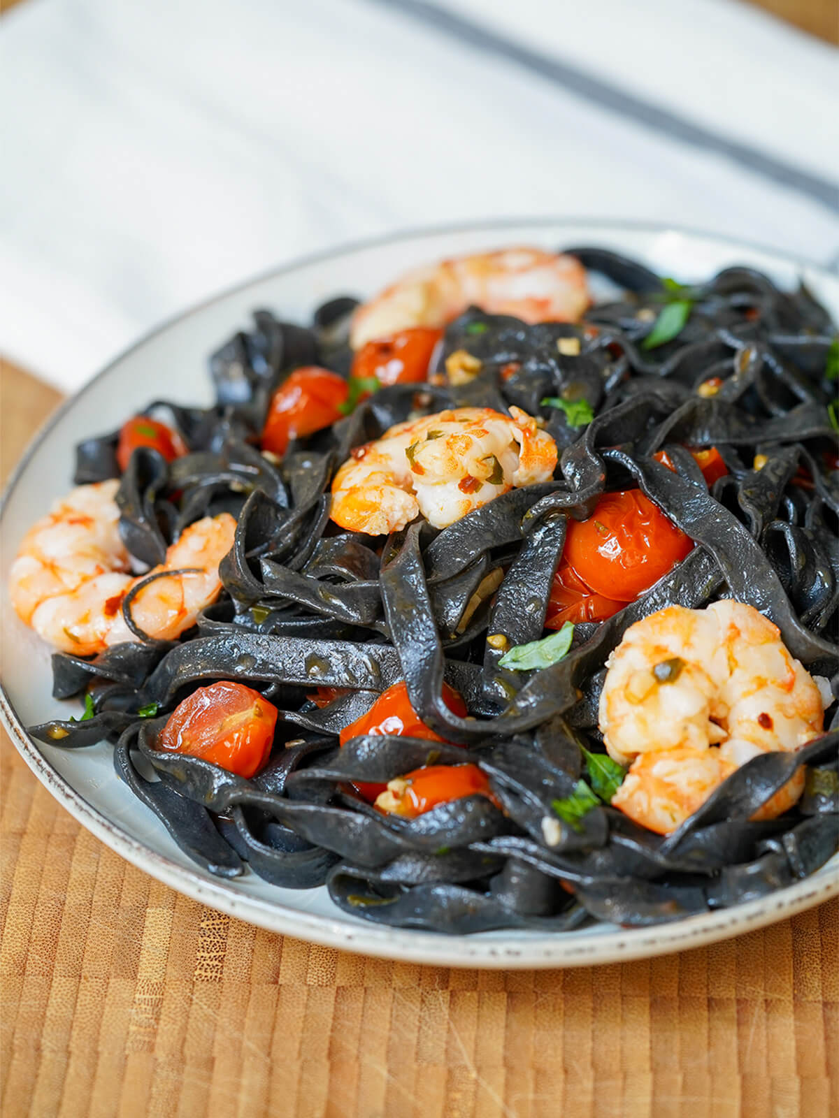 partial view of a plate of squid ink pasta with shrimp and tomato with cloth in background.