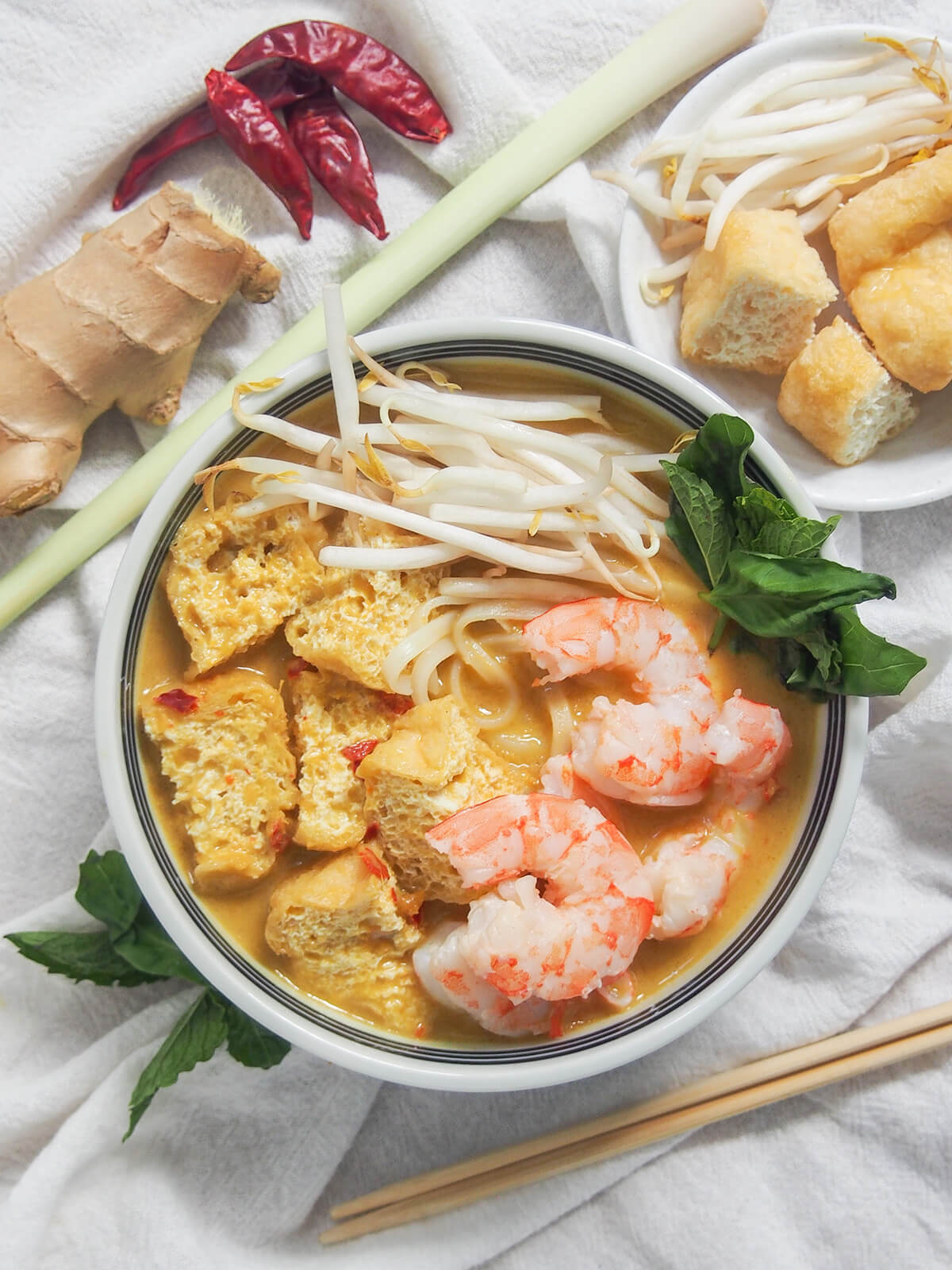 overhead view of a bowl of Singapore laksa with additional herbs and toppings around bowl.