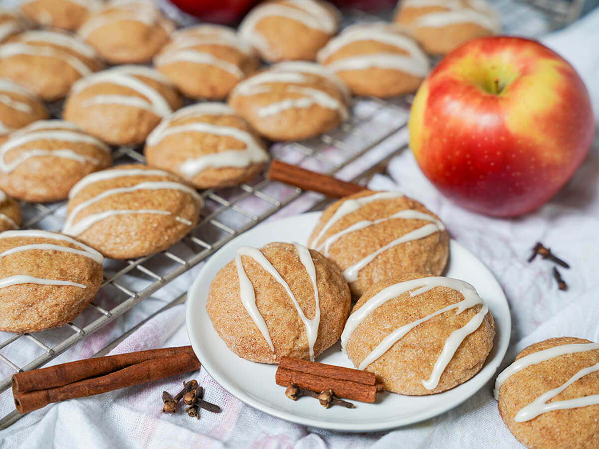 plate of three apple cider cookies with more on cooling rack behind and spices and apple around.