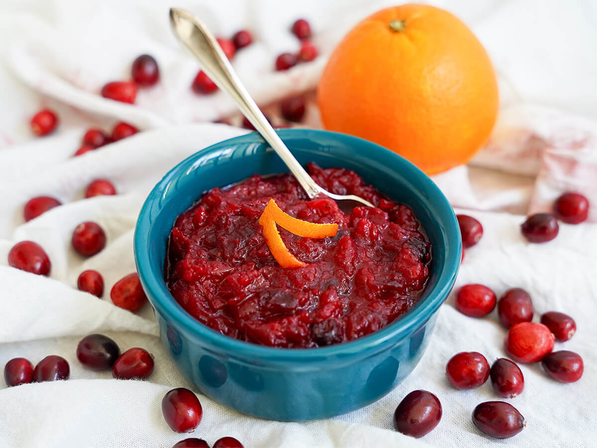 dish of cranberry sauce with orange with a spoon in back of bowl, orange behind and cranberries around.