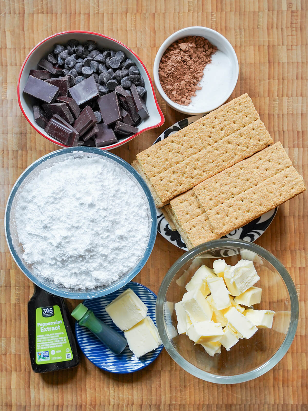 bowls of confectioner's sugar, chocolate, graham crackers, butter, cocoa powder with peppermint and green color bottles below.