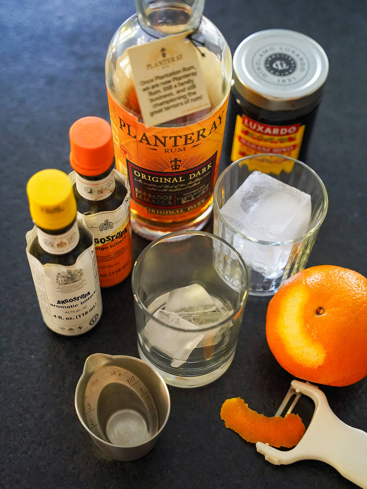 two glasses with ice, bottle of rum, bottles of bitters and an orange with a strip peeled from it.