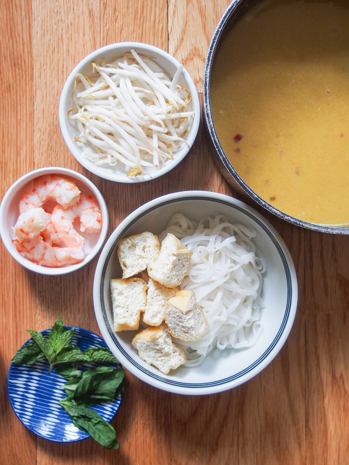 putting together bowl of Singapore laksa bowl of noodles and tofu puffs with beansprouts and shrimp to side, pot of soup base to other side.