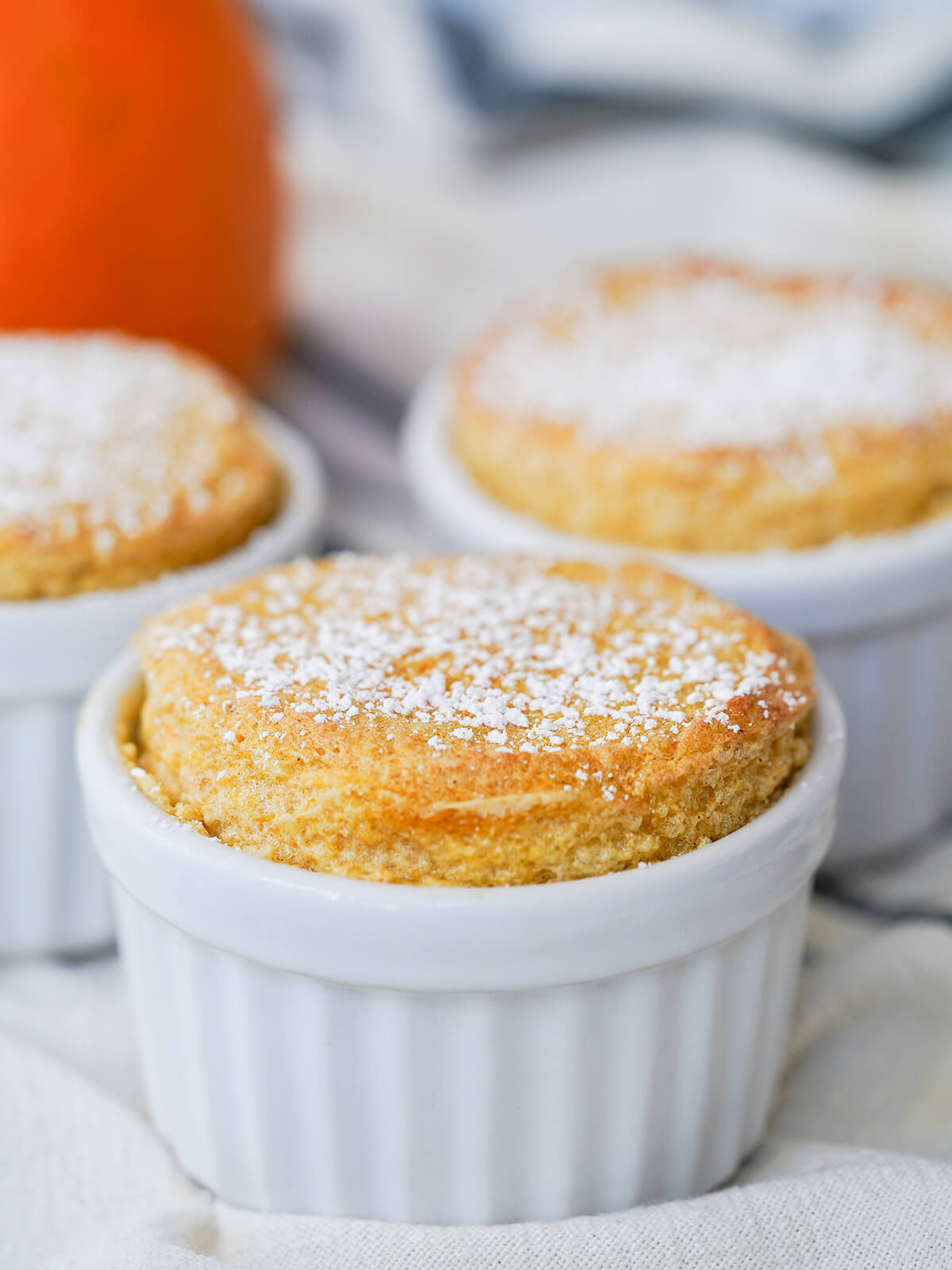 This pumpkin souffle is a light, gluten free dessert full of the flavors of pumpkin pie. It's gently spiced, with a luscious sweetness and texture. Delicious any excuse. side view of a pumpkin souffle with others blurred behind.