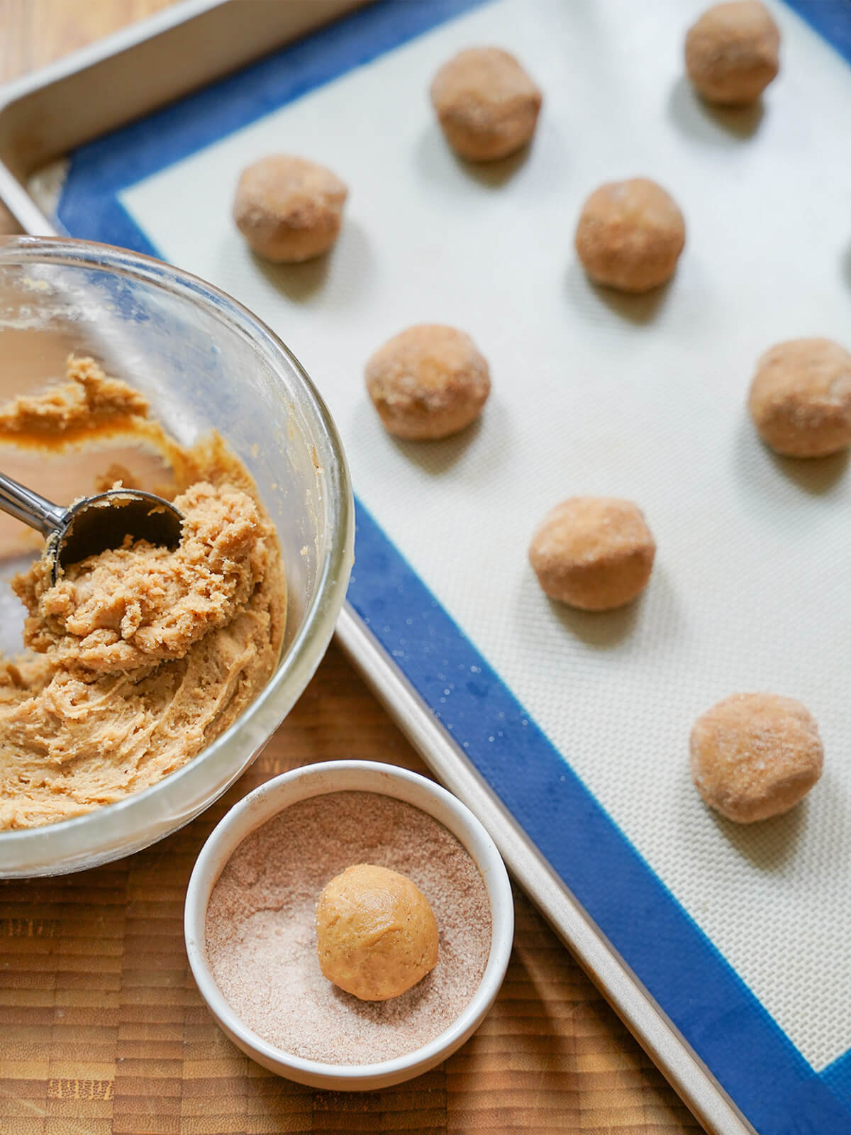 bowls of dough with scoop in it, a ball of dough in dish with cinnamon sugar and finished balls on lined baking sheet.