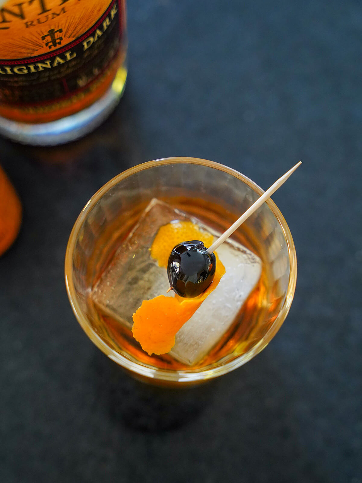 overhead view of glass with rum old fashioned over large square ice cube with twist of orange and cherry on top.
