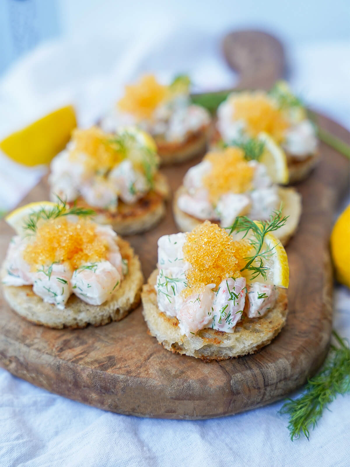six pieces of toast Skagen on a wooden board with lemon and dill at edges.