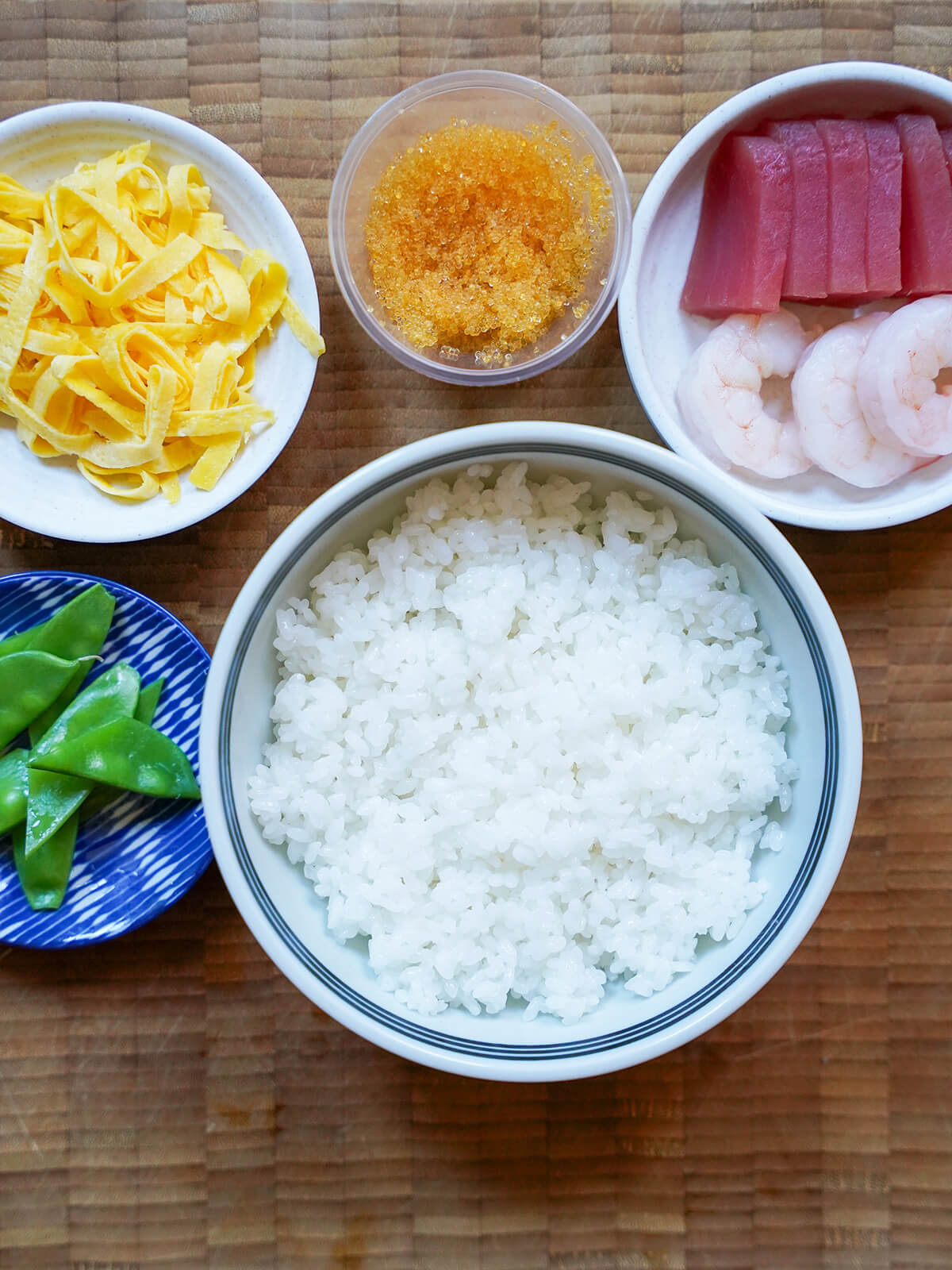 bowl of rice with dishes with tuna sashimi, shrimp, shredded omelette and fish roe above.