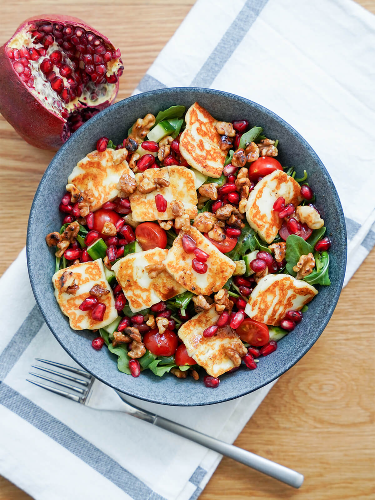 This halloumi pomegranate salad uses a short list of ingredients but is packed with flavor, color and texture. It's bright, versatile and delicious. overhead view of a bowl of halloumi pomegranate salad with pomegranate to side, fork below bowl.