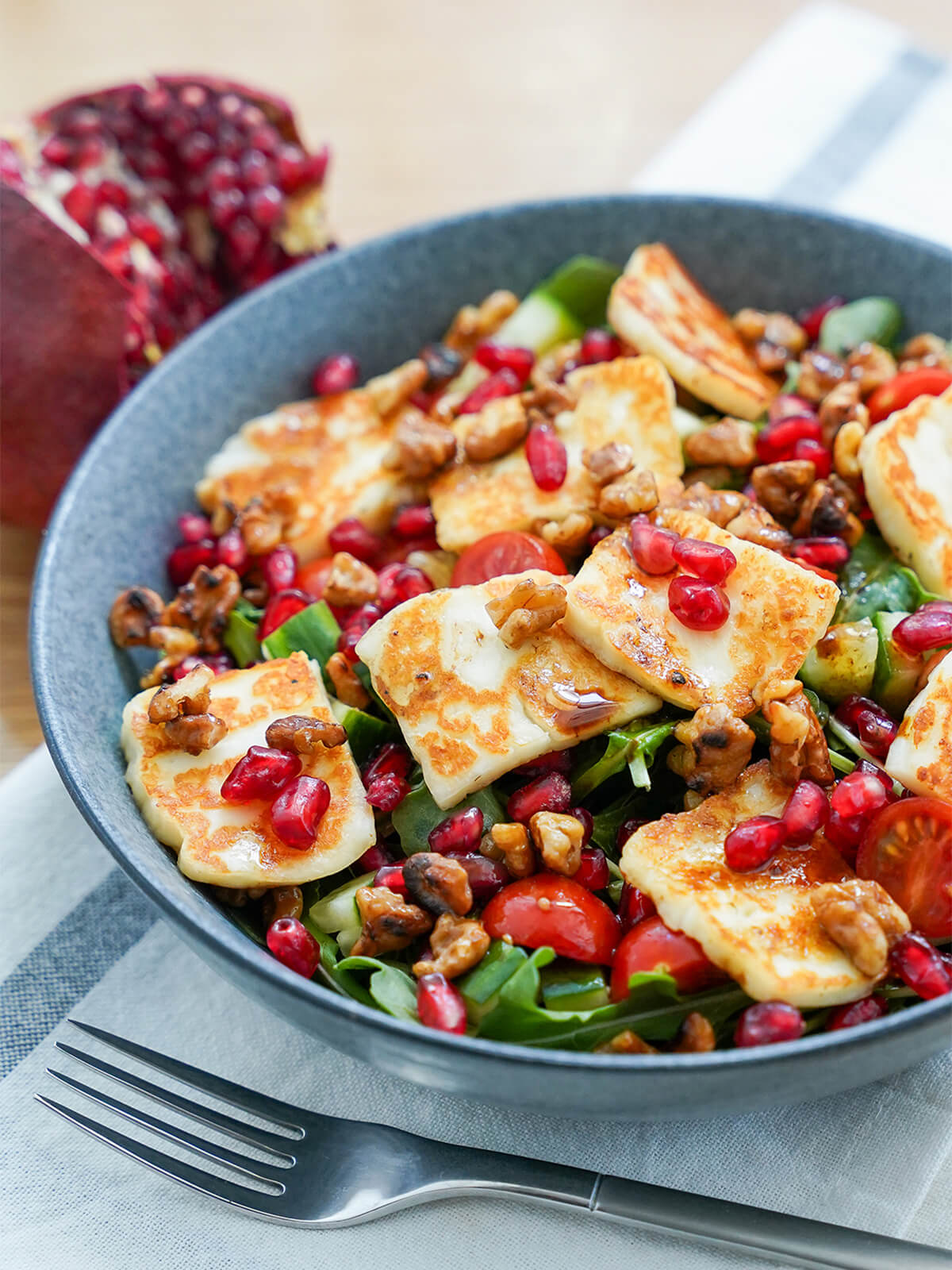 This halloumi pomegranate salad uses a short list of ingredients but is packed with flavor, color and texture. It's bright, versatile and delicious. side view of a bowl of halloumi pomegranate salad with pomegranate behind bowl and fork in front.