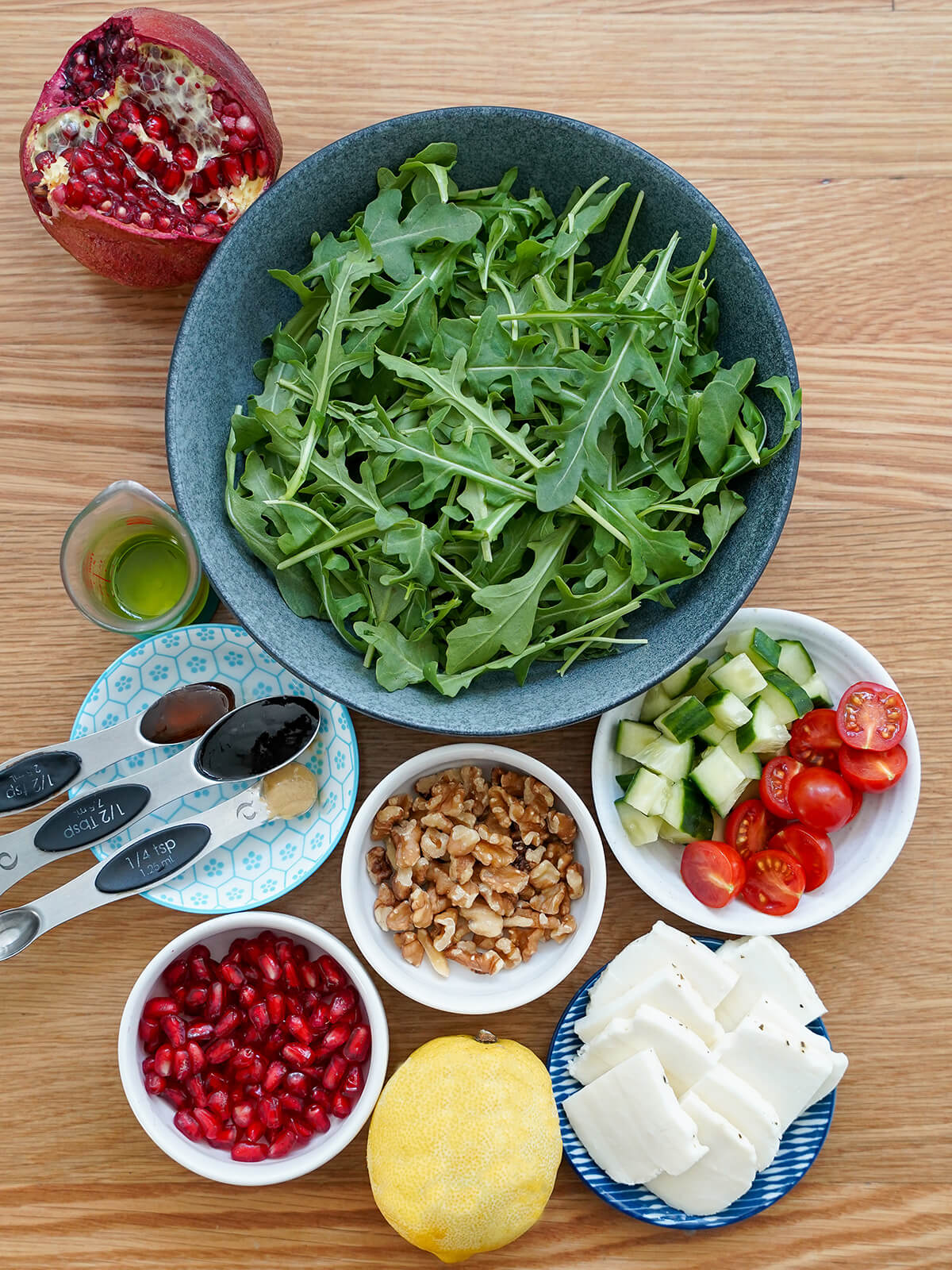 ingredients for halloumi pomegranate salad bowl of arugula, small bowls of cucumber, tomatoes, walnuts, pomegranate, halloumi slices and lemon and spoons with liquids to side.