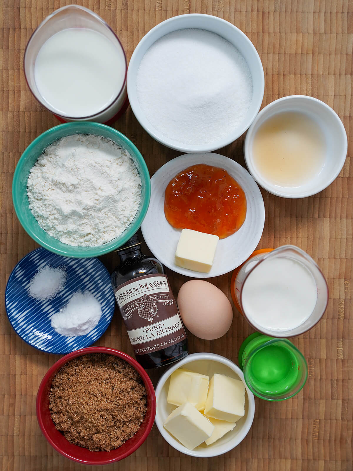 bowls with sugars, flour, butter, measures with milk, cream and dish with apricot jam, bottle of vanilla and egg in middle.